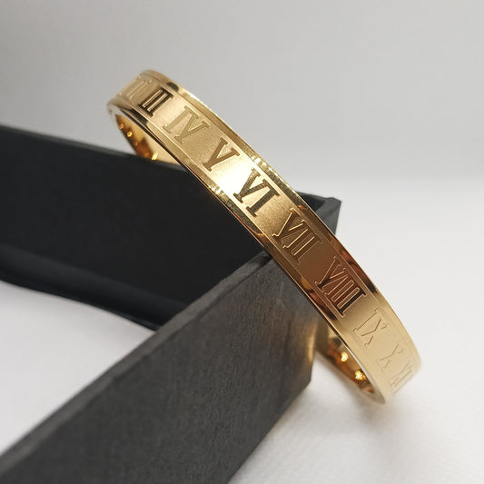 Gold bracelet with Roman numeral design on a black stand against a white background